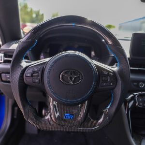 Toyota GR Supra custom carbon fiber steering wheel installed, black perforated leather, blue stitching, leather airbag cover, gloss carbon